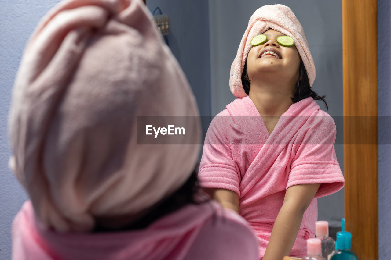 A cute indian girl child in pink bathrobe applying cucumber slices to her eyes in front of mirror