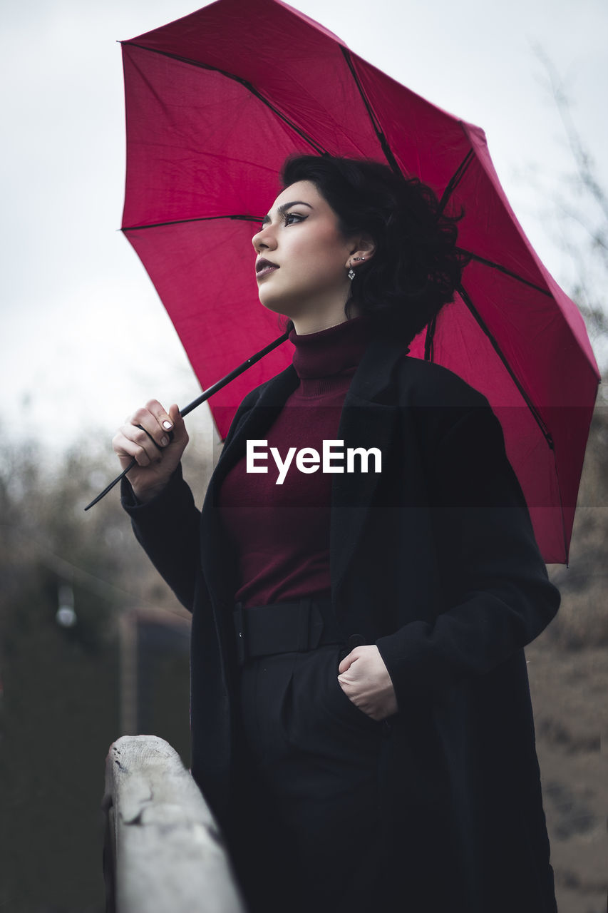 Woman looking away holding umbrella standing against sky