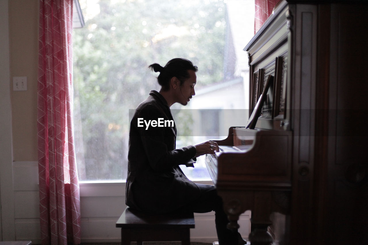 Side view of man playing piano against window at home