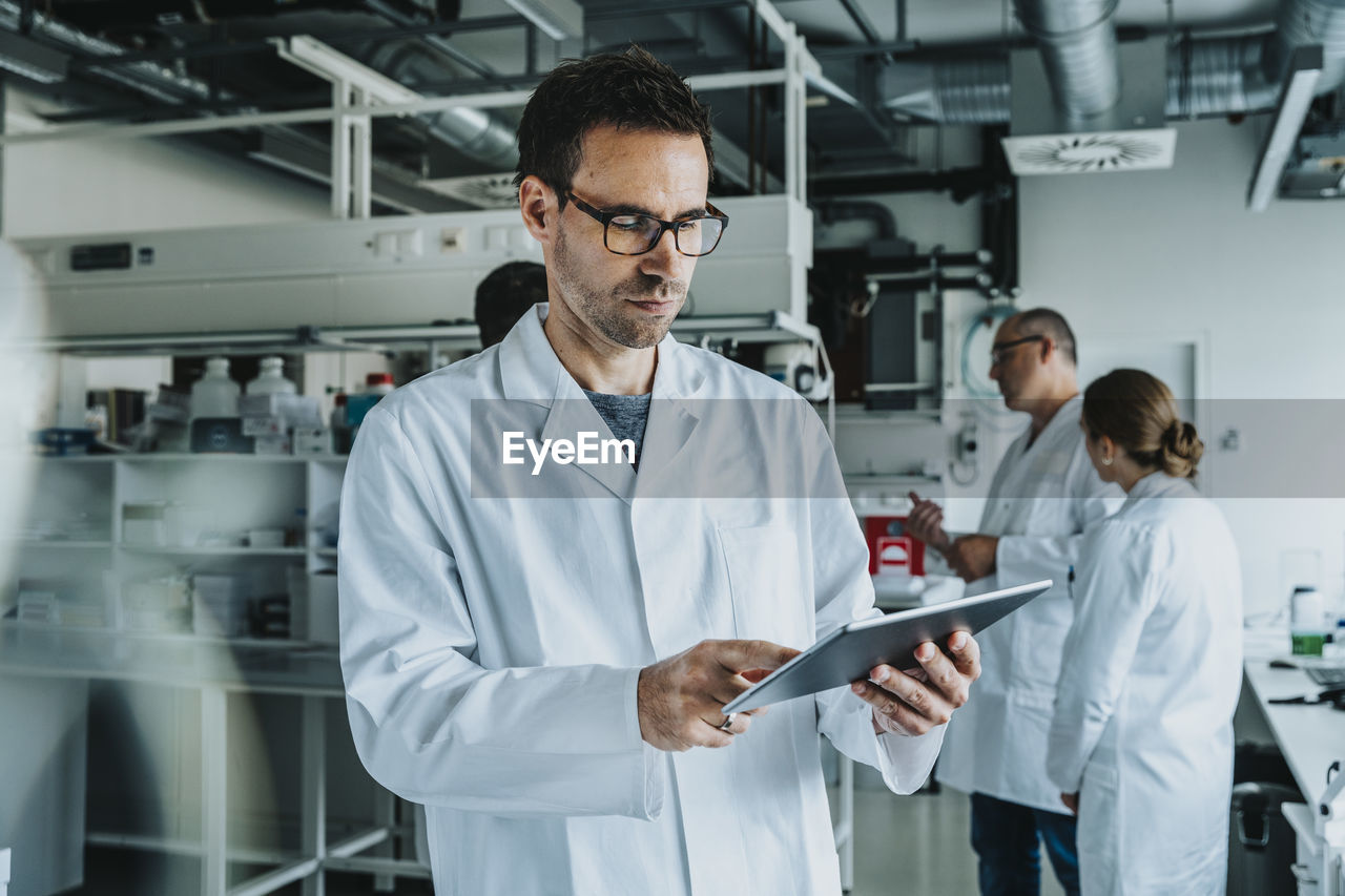 Scientist wearing eyeglasses using digital tablet while standing with coworker in background at laboratory