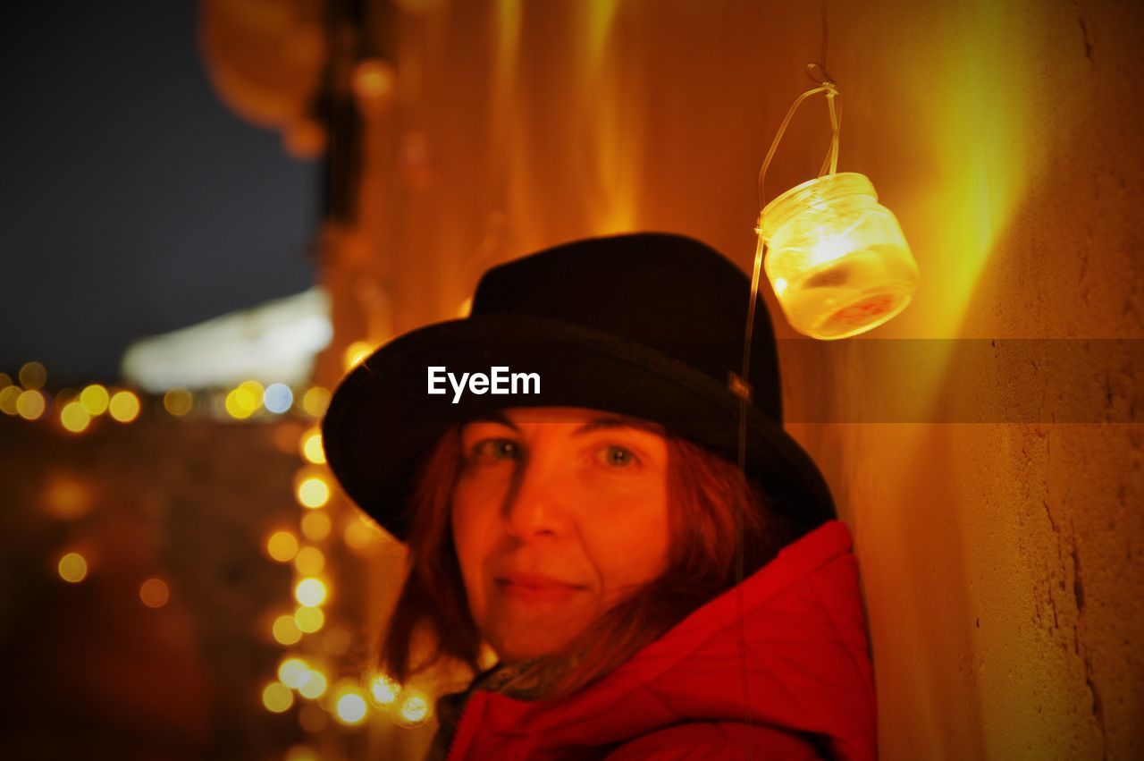 Portrait of woman wearing hat against illuminated light at night