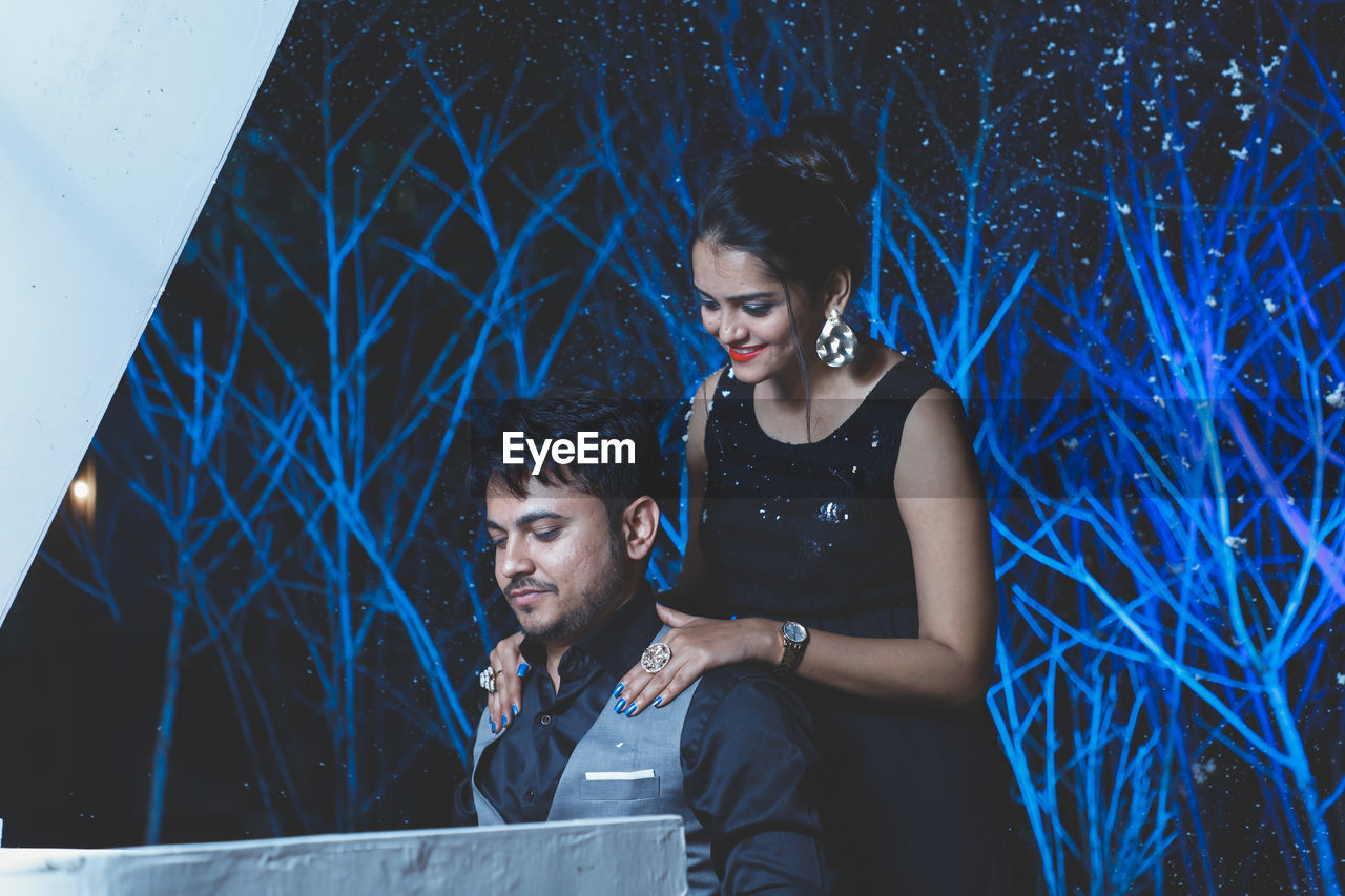 Young couple looking at camera at night against blue bushy background