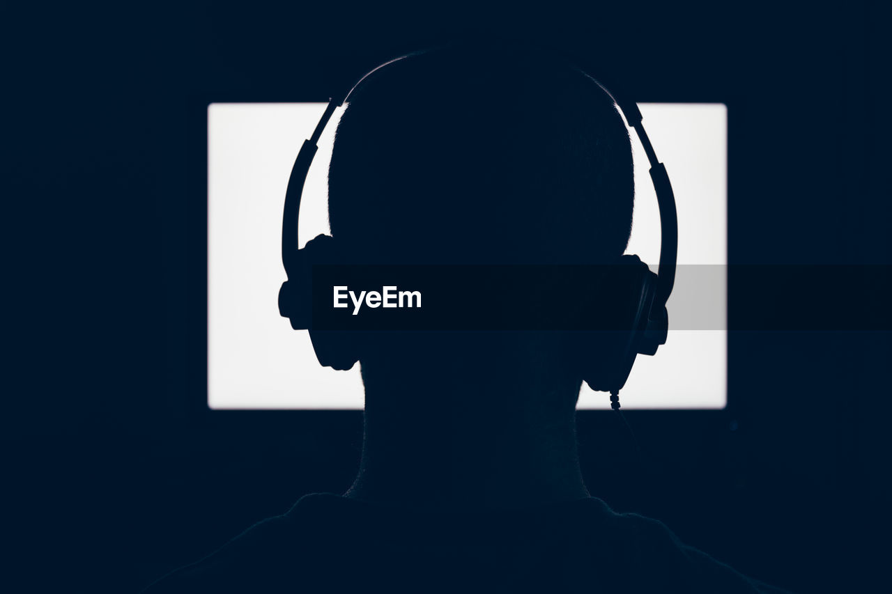 Close-up of silhouette man wearing headphones against illuminated device screen in dark