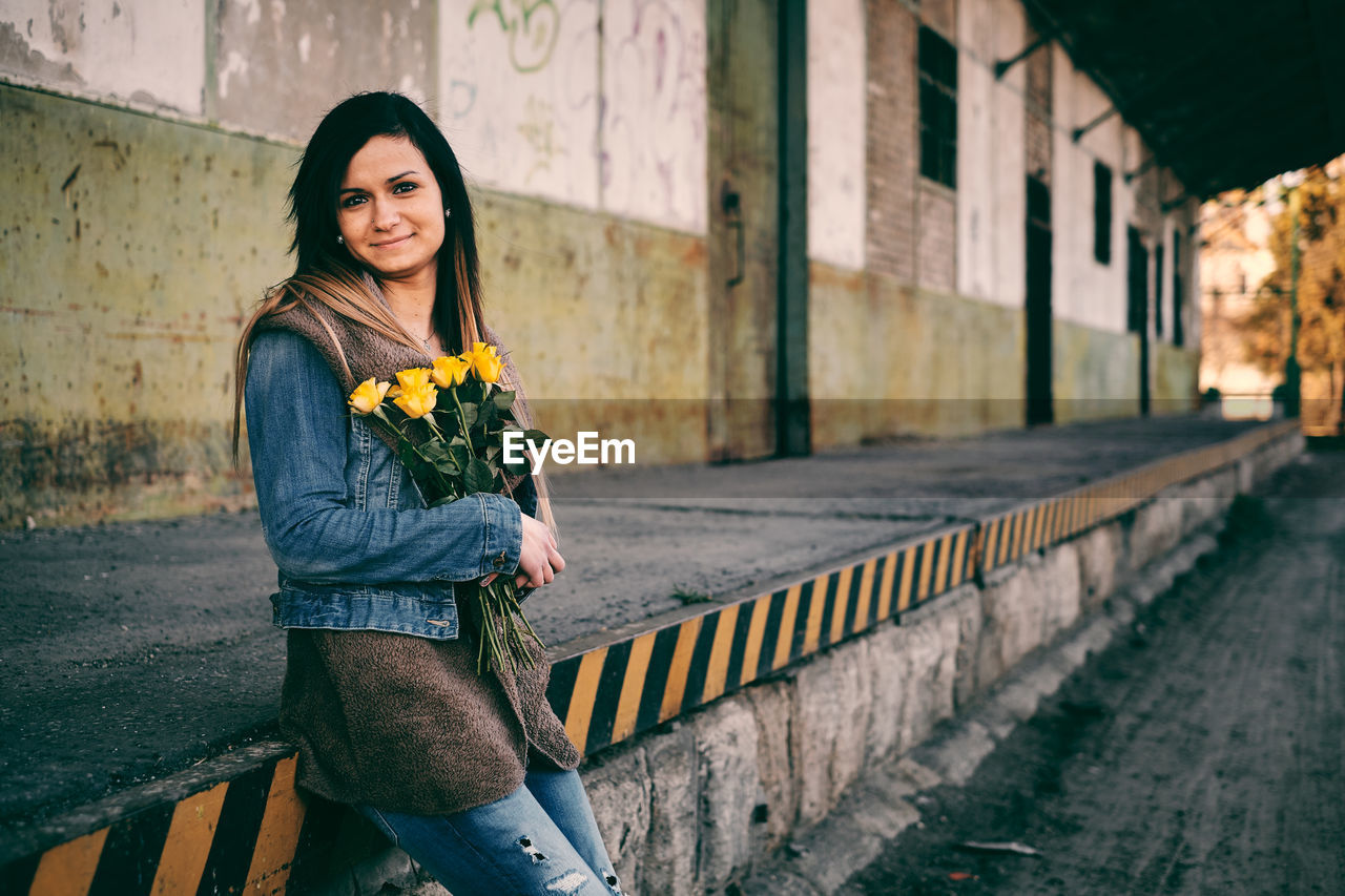 Portrait of smiling young woman holding roses while leaning on retaining wall