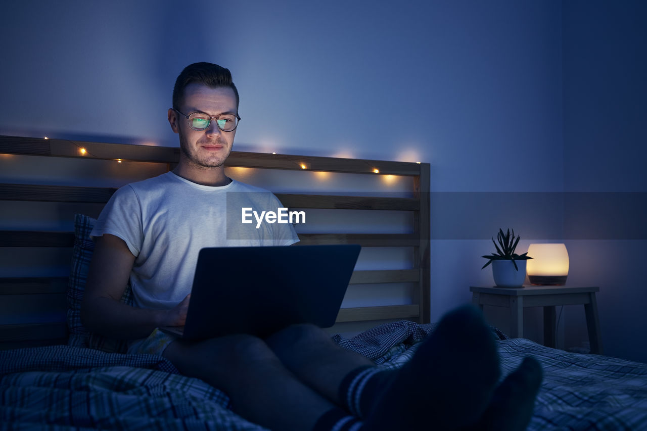Man with eyeglasses sitting on bed in bedroom and using laptop. 