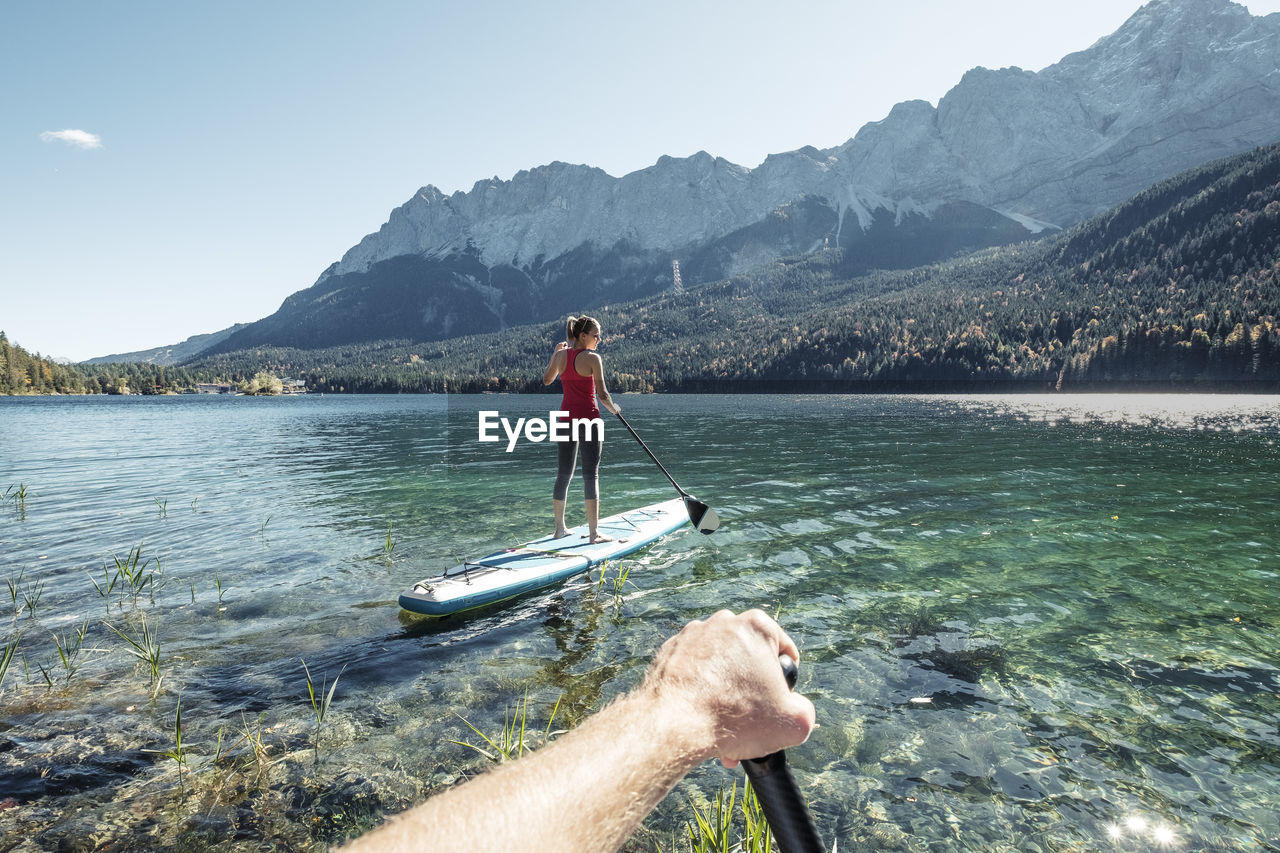 Germany, bavaria, garmisch partenkirchen, young couple stand up padling on lake eibsee, overlooking zugspitze mountain