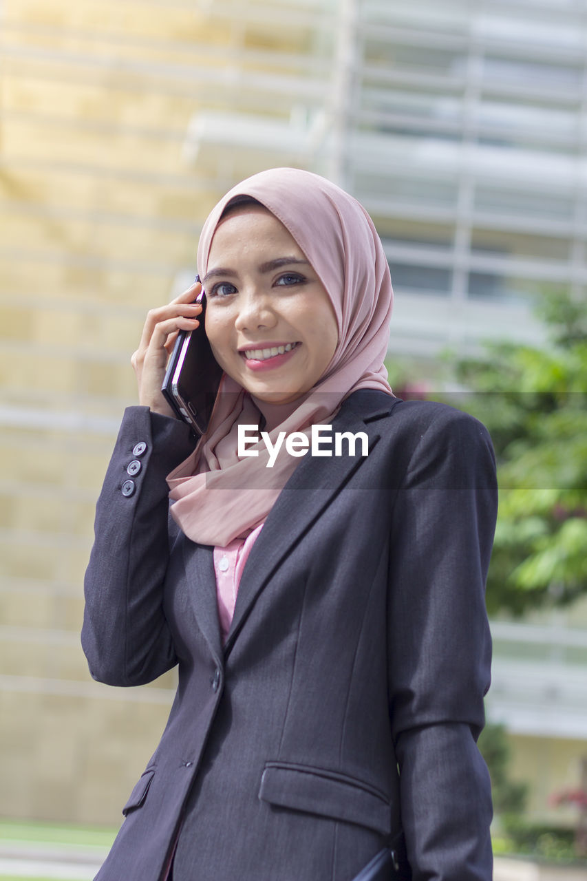 Low angle portrait of happy businesswoman using smart phone while standing against office building