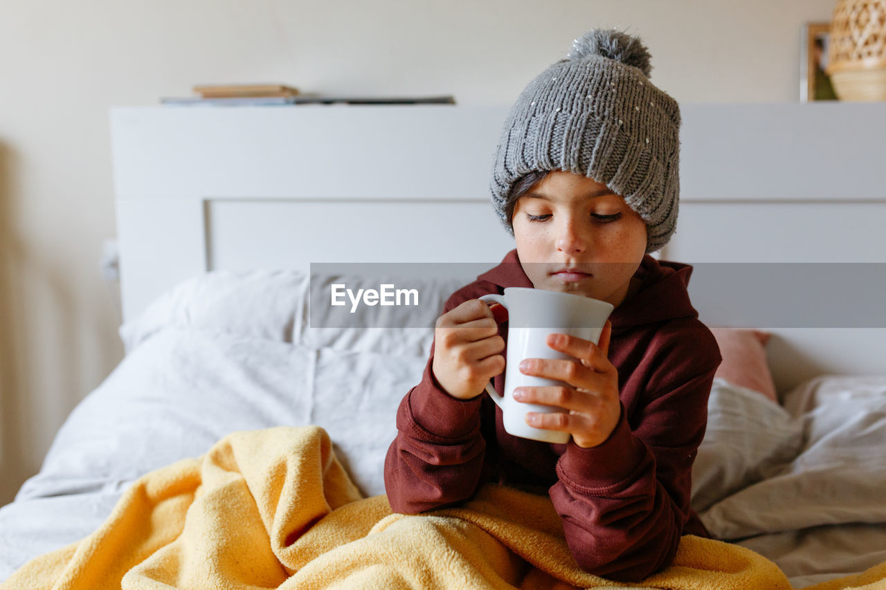 Child wearing grey warm hat sits on the bed under warm blanket and drinks hot tea