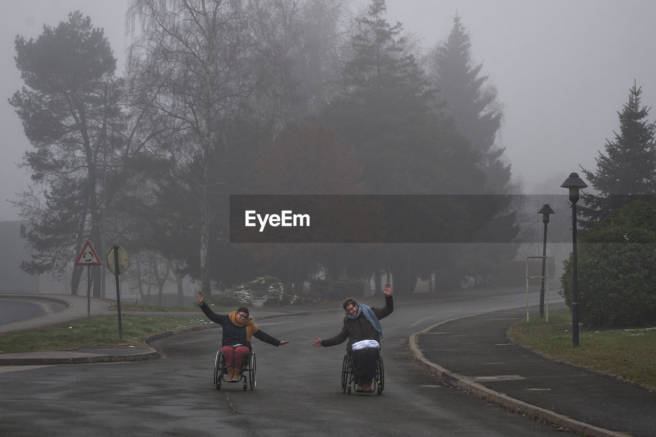 Friends with arms outstretched riding on wheelchairs in park during winter