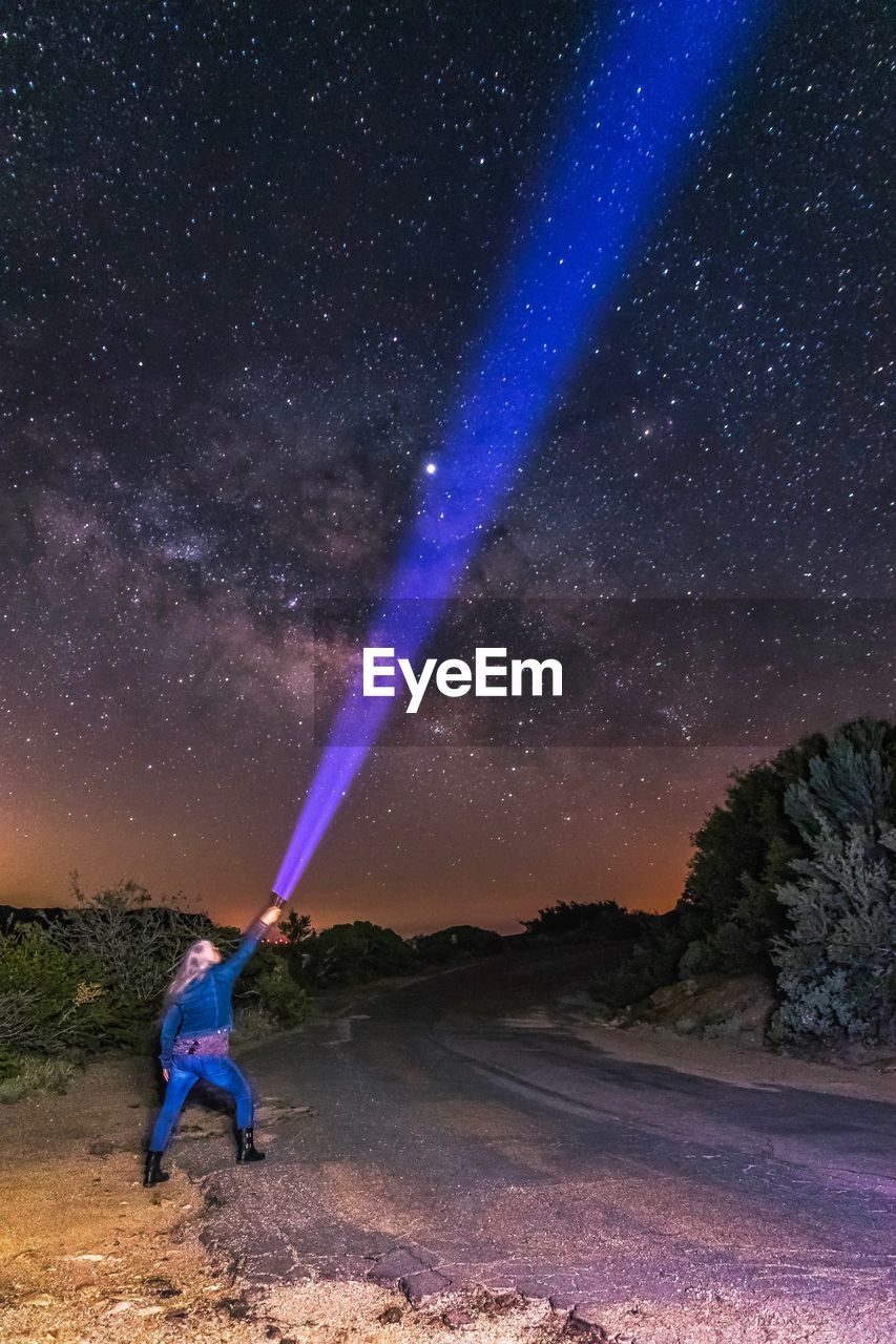 Woman holding flashlight towards star field in field against sky at night