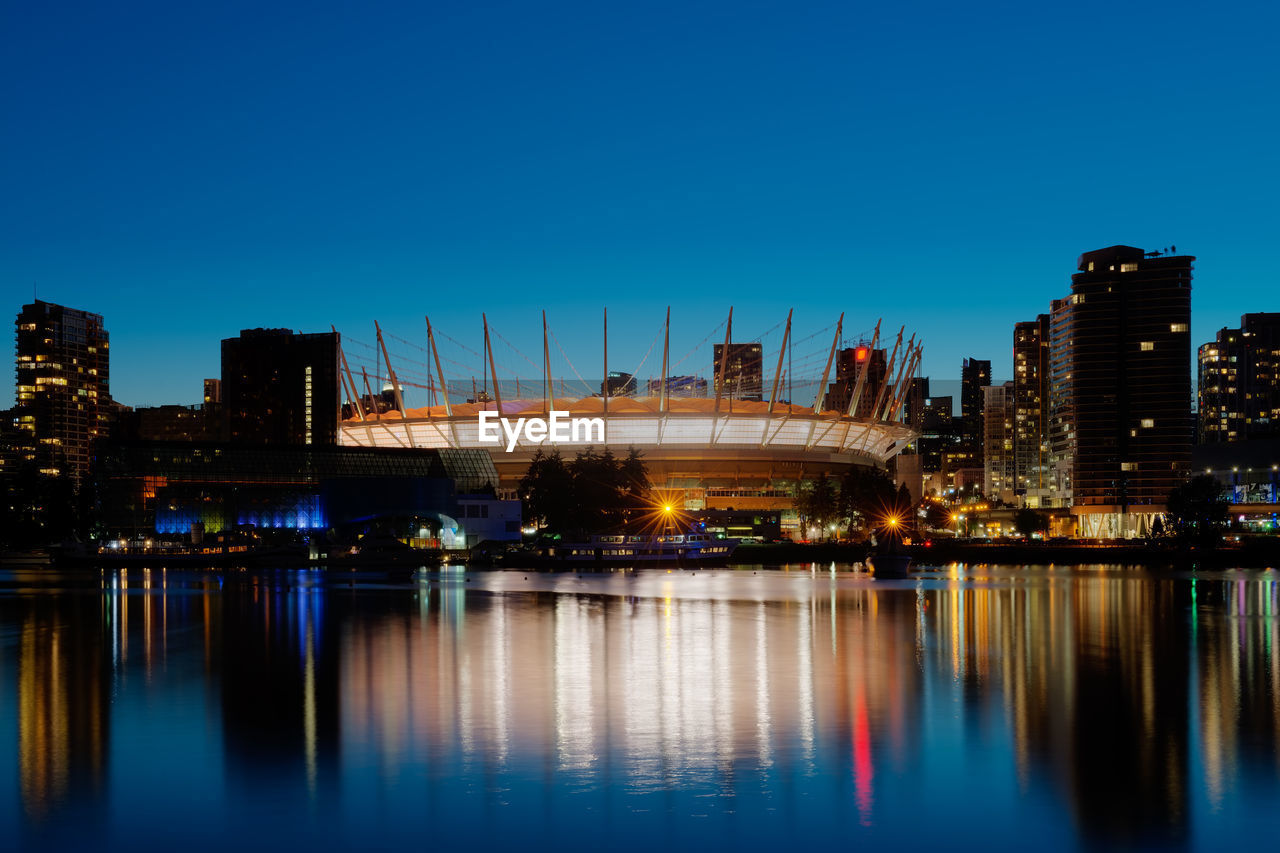 ILLUMINATED CITYSCAPE BY RIVER AGAINST CLEAR BLUE SKY