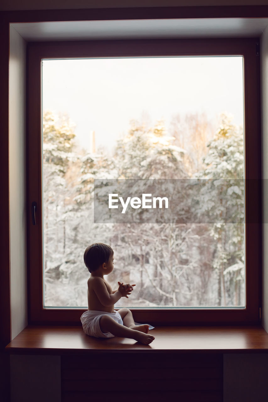 Baby boy in bath towel after washing sitting on the windowsill at the big window in winter