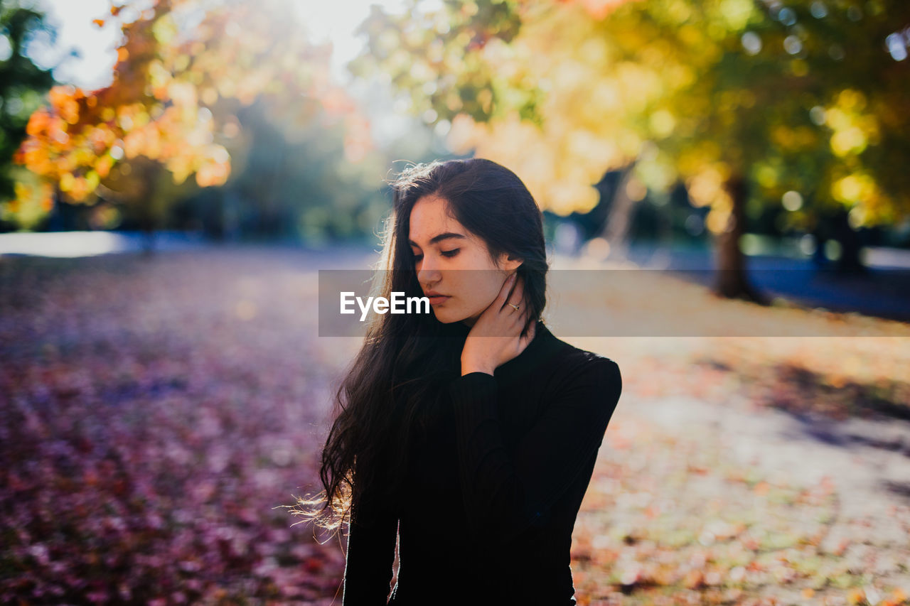 Young woman looking down while standing against trees in forest during autumn 