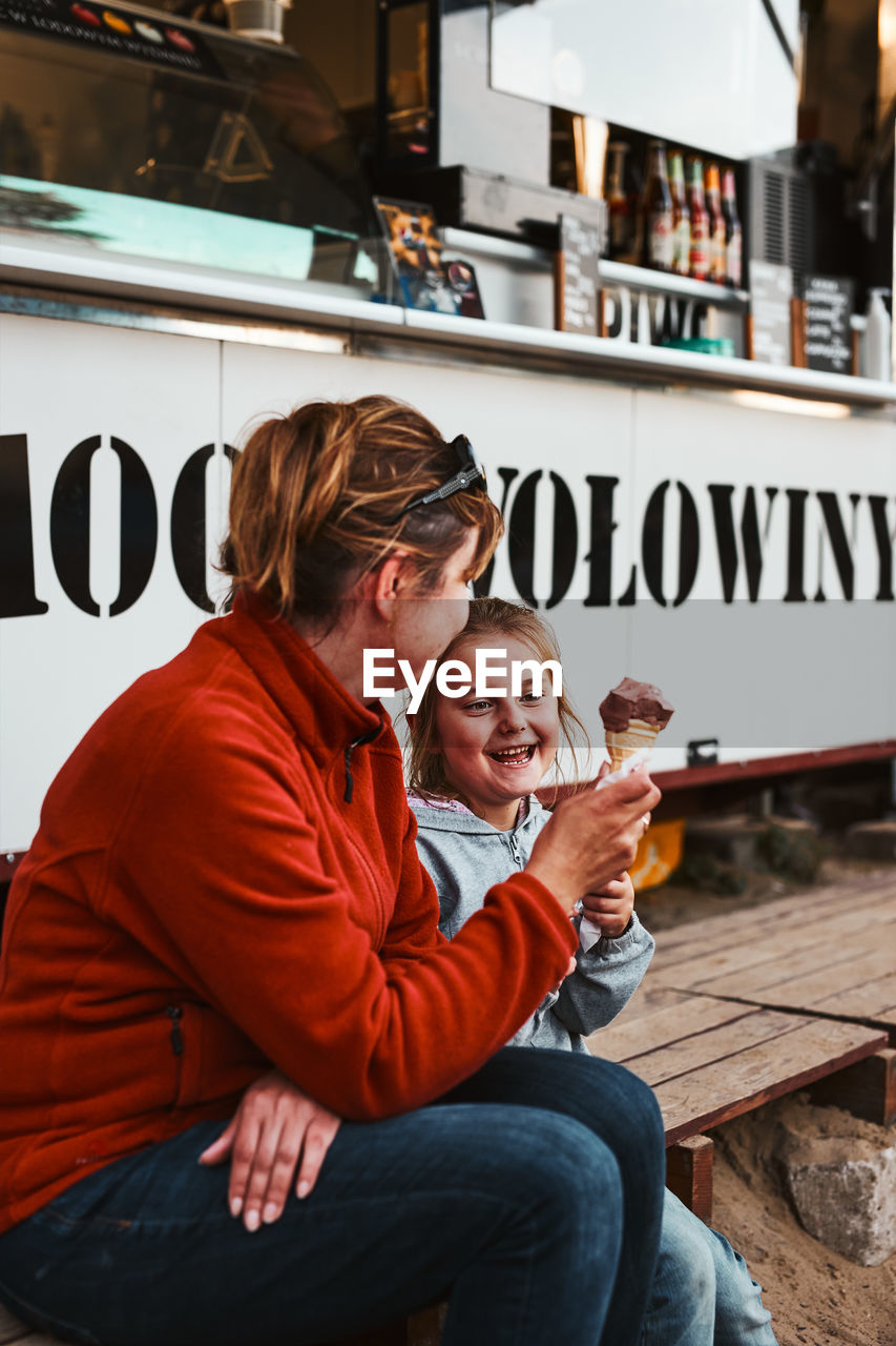 Mother and her daughter eating ice cream sitting on a step in front of food truck during vacations