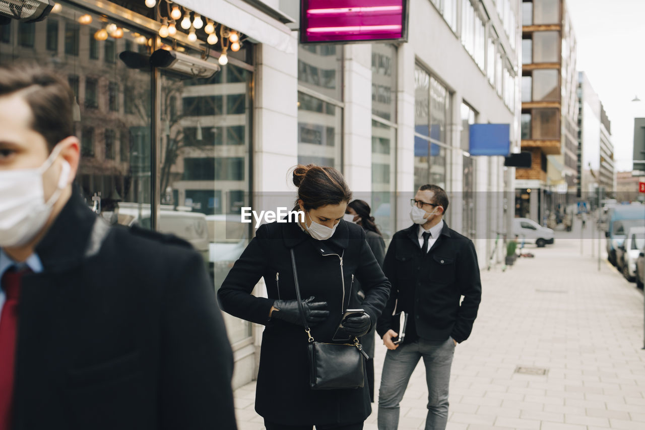 Female entrepreneur holding smart phone while standing in line with colleagues during covid-19