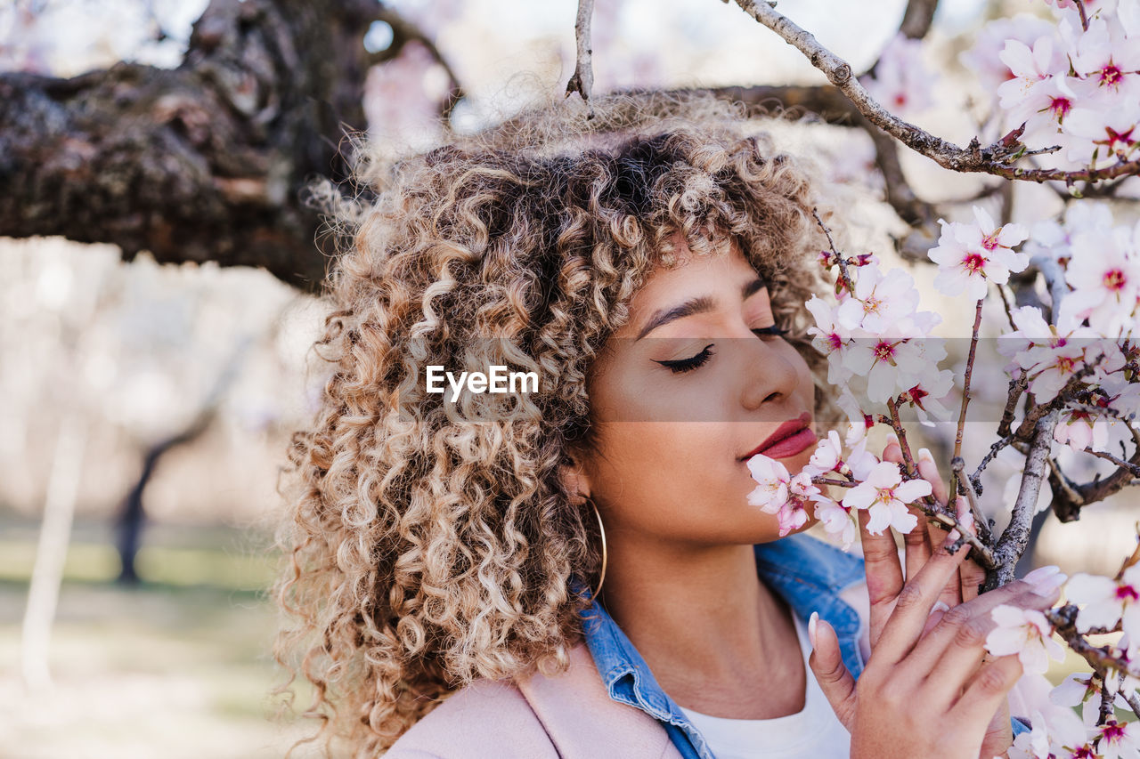 Portrait of beautiful hispanic woman with afro hair in spring smelling pink blossom flowers. nature