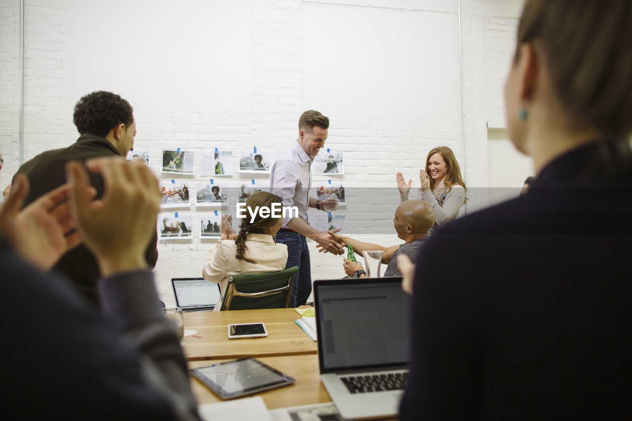 Business people applauding for businessman giving handshake to male colleague in board room