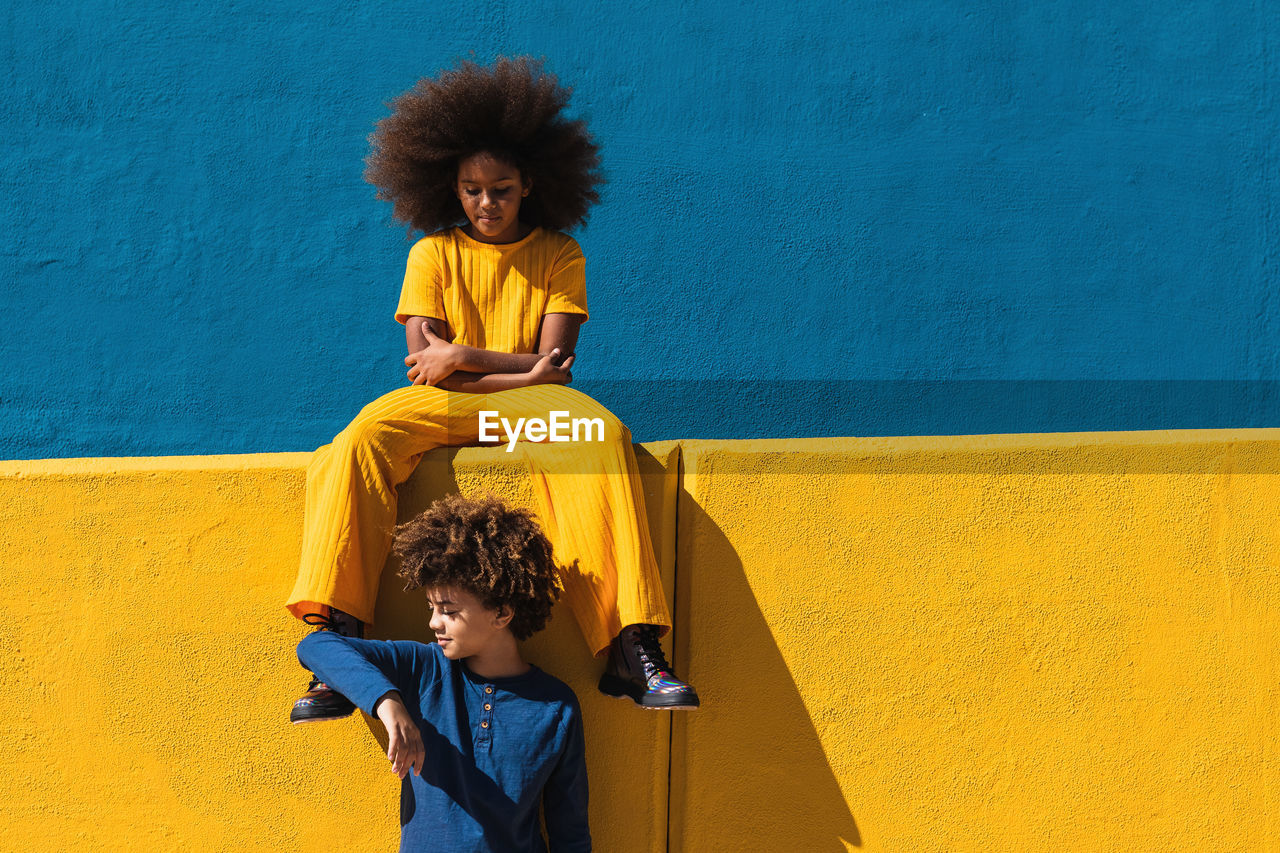 Teen african american girl and boy with afro hairstyle wearing colorful clothes resting together against blue and yellow wall in sunny day on street