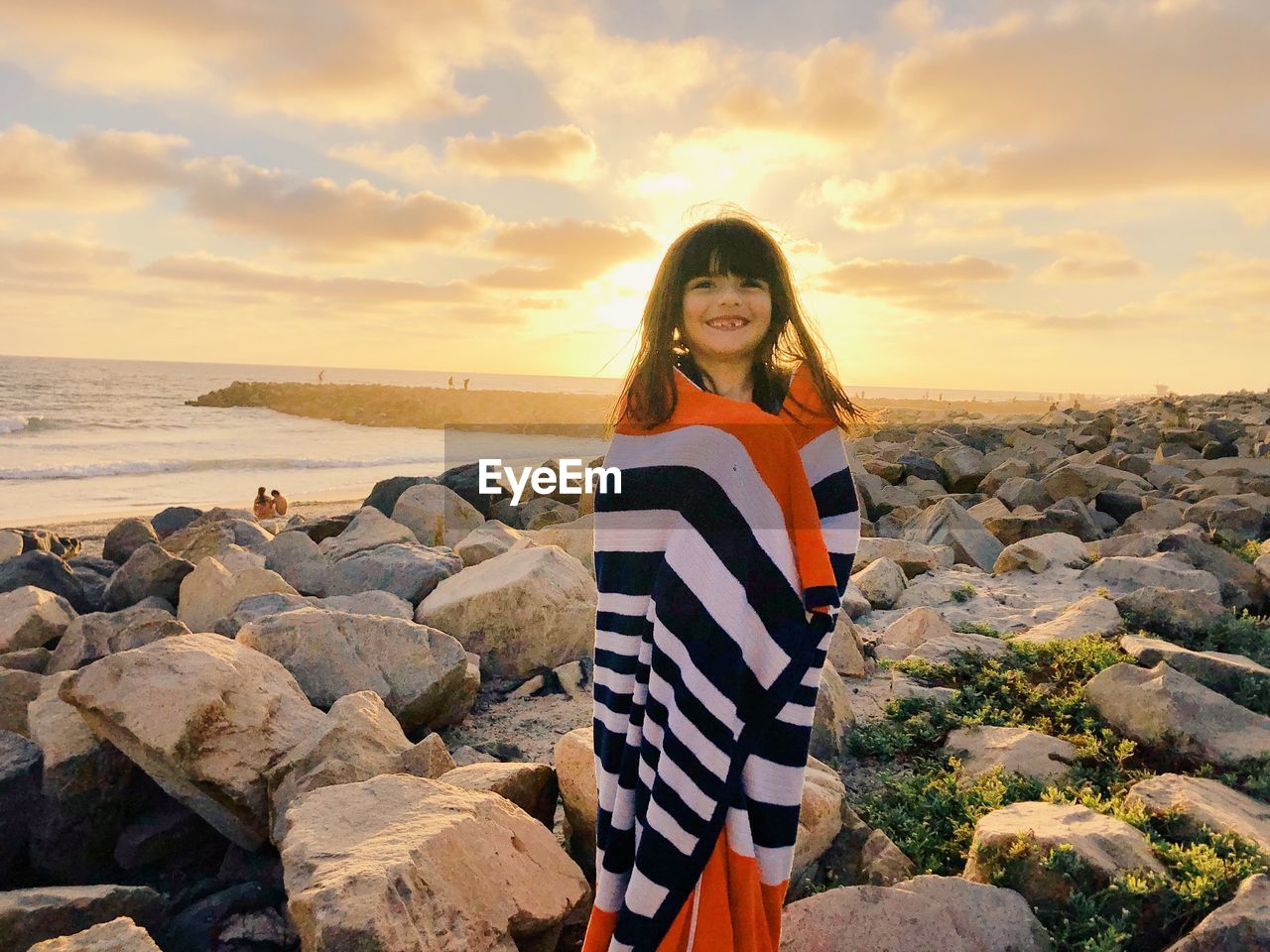 Smiling girl wrapped in towel while standing on rocks at beach against sky during sunset