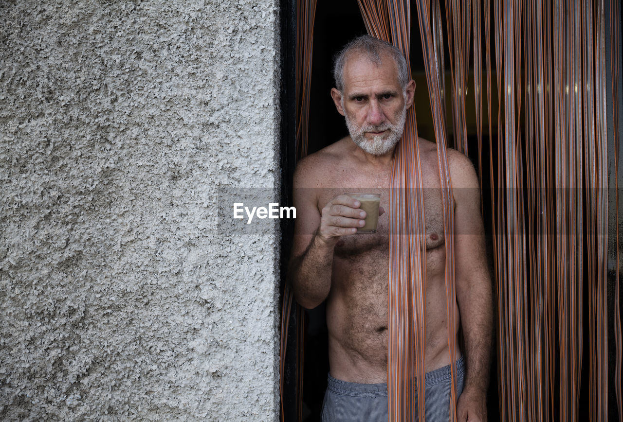 Portrait of shirtless man having coffee on door standing against wall