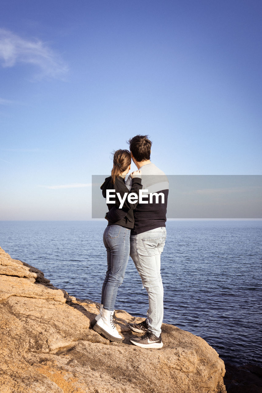 sky, sea, water, two people, adult, men, full length, nature, togetherness, standing, rock, casual clothing, horizon over water, love, positive emotion, beach, women, horizon, emotion, land, ocean, blue, vacation, young adult, leisure activity, trip, holiday, scenics - nature, beauty in nature, copy space, romance, bonding, day, embracing, clear sky, tranquility, rear view, coast, relaxation, looking at view, travel, outdoors, lifestyles, happiness, shore, sunlight, tranquil scene, looking, female, travel destinations, sunny, affectionate