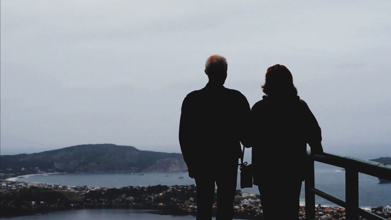 SILHOUETTE PEOPLE STANDING BY RAILING AGAINST SEA AND MOUNTAINS