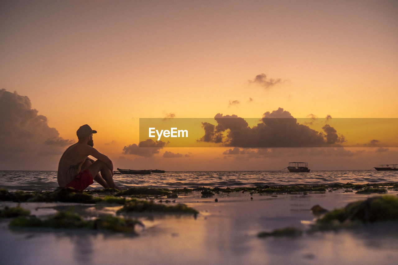 Man sitting at beach against sky during sunset