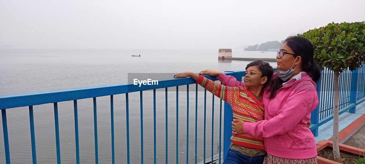 Mother and son embracing while standing by railing against sea