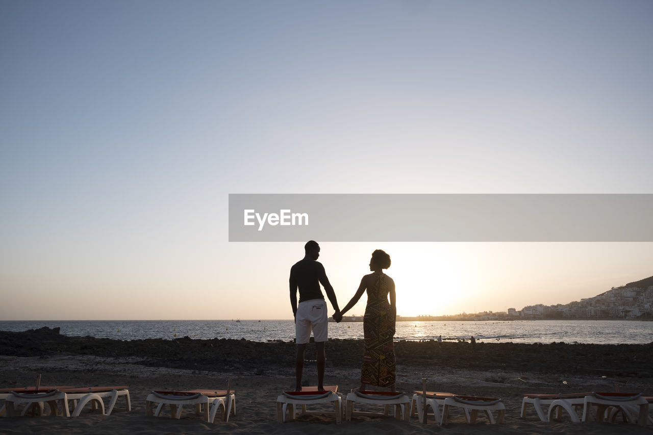 Couple standing at beach against sky during sunset