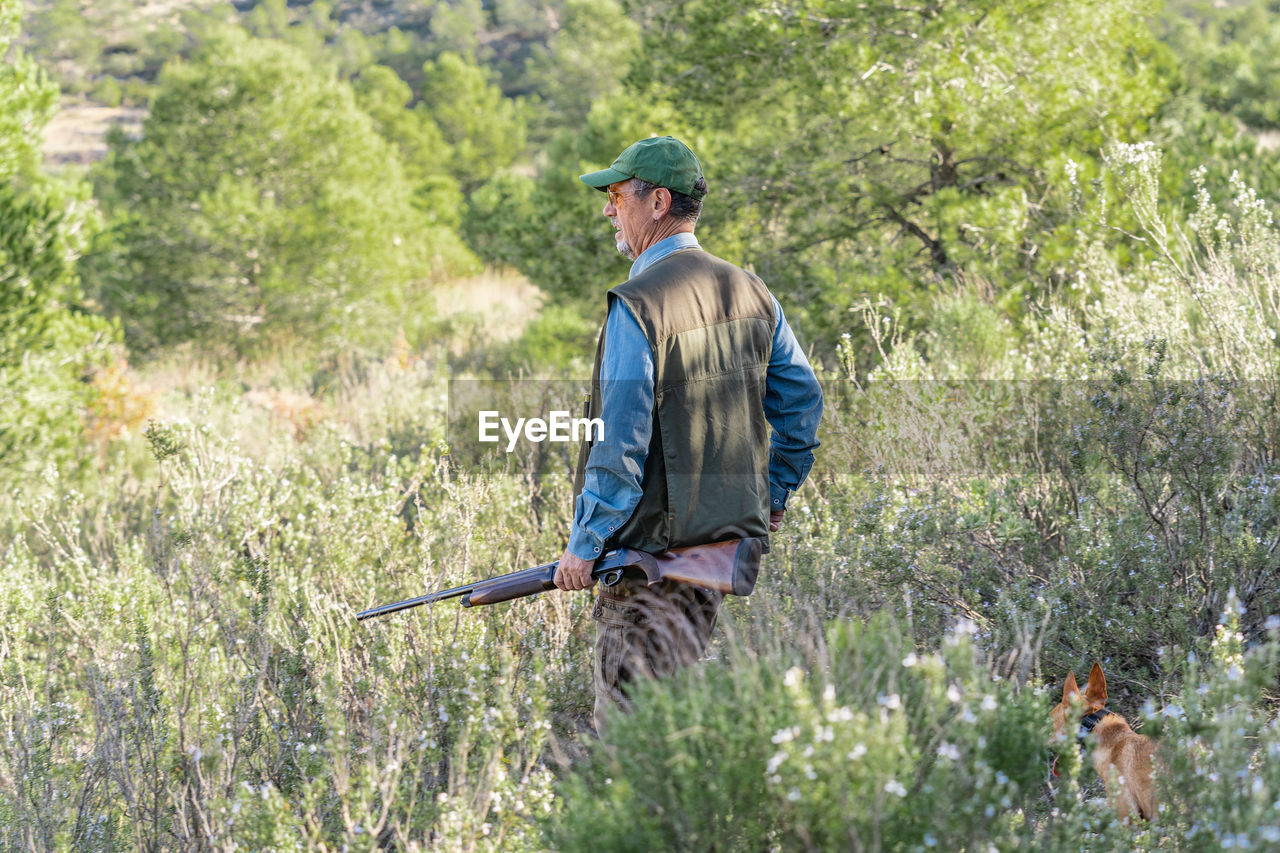 Side view of male hunter in special hunting vest and green cap holding rifle and looking away among tall grass