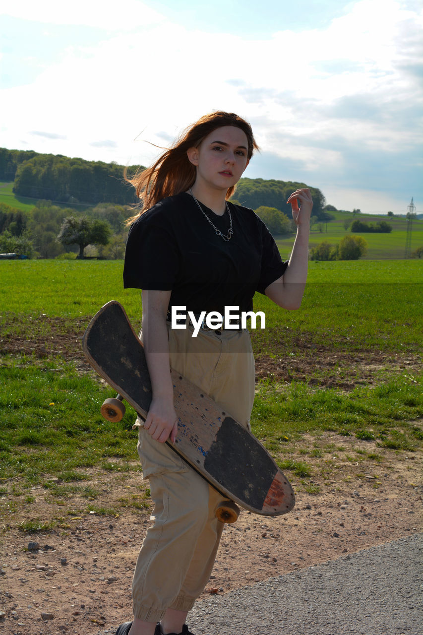 Young girl with a skateboard in her hand, stands in green nature, one arm up