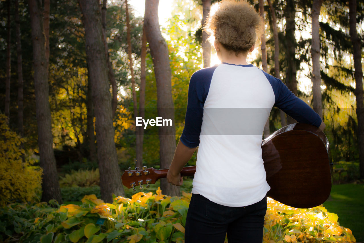 Rear view of boy holding guitar against trees at park