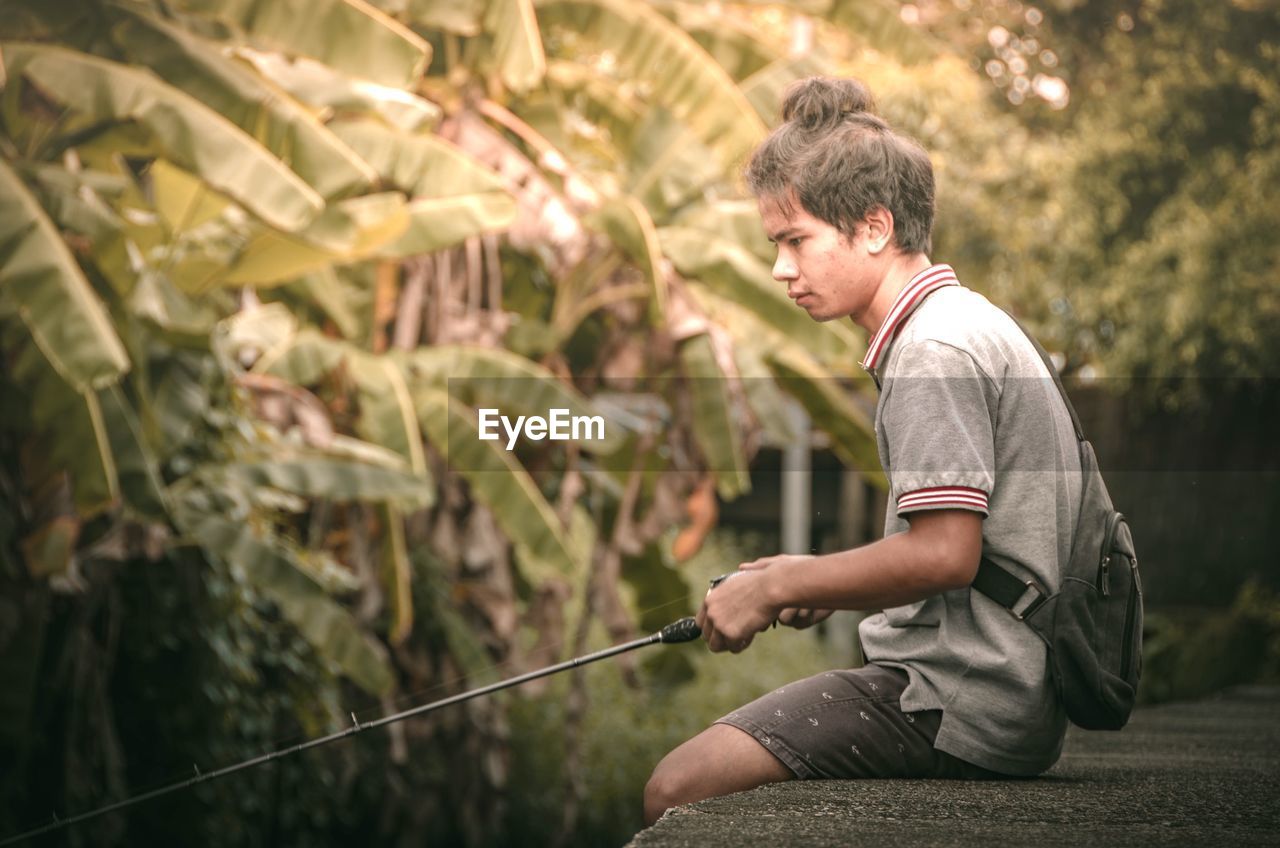 Side view of young man with fishing rod sitting in forest