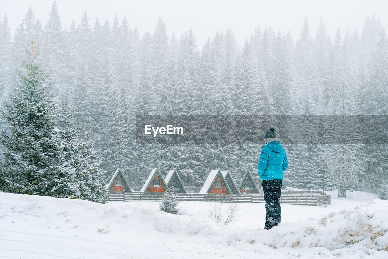 Man in worm clothing looking at wooden cabins during blizzard