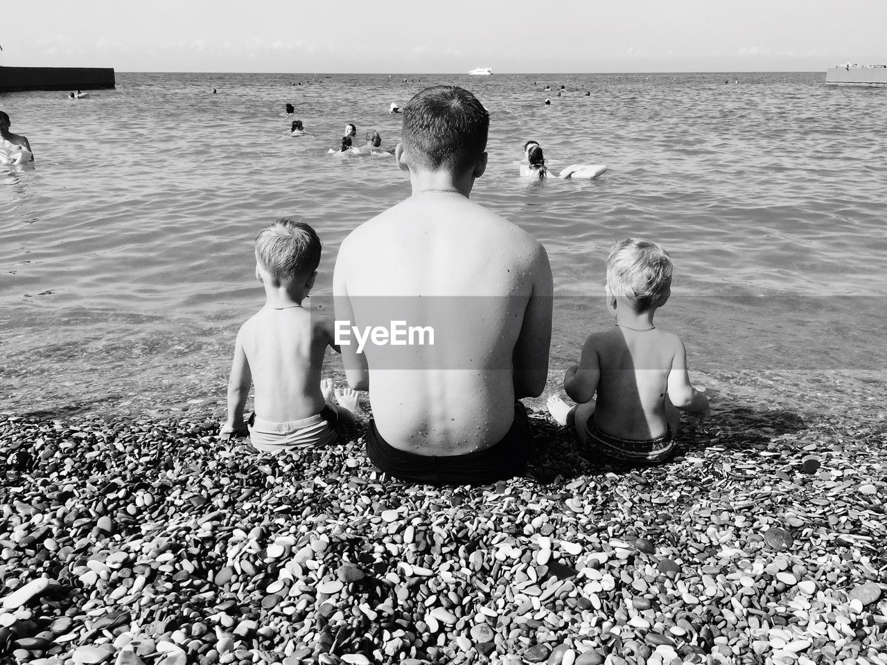 Rear view of father and sons sitting on sea shore against sky