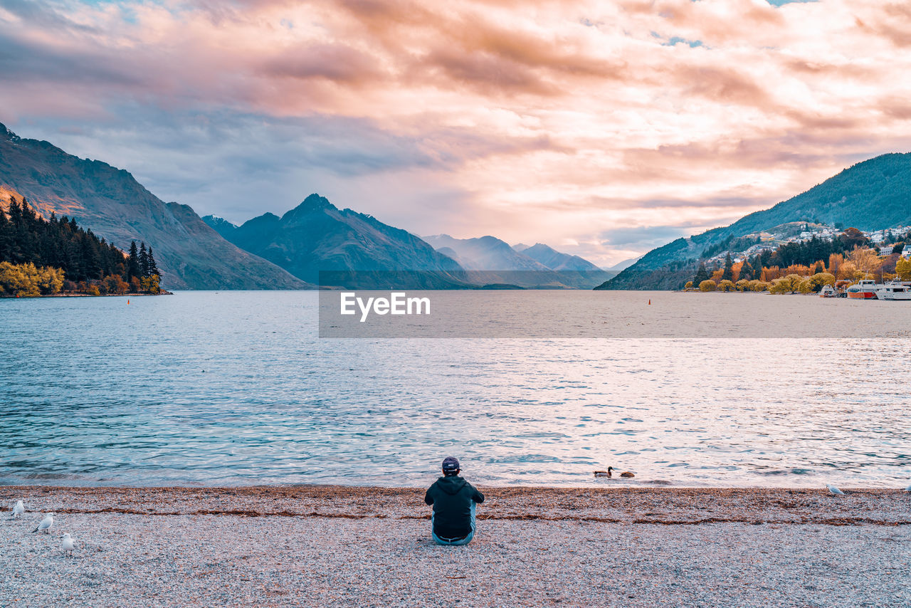 Rear view of woman sitting on land by lake against mountains and sky