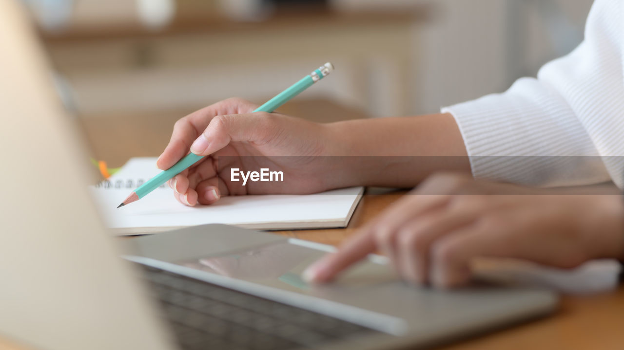 Cropped hands of woman using laptop while writing in diary