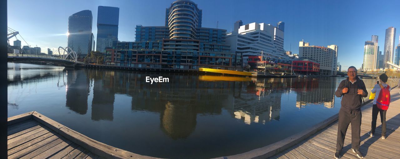 REFLECTION OF WOMAN STANDING ON RIVER BY BUILDINGS AGAINST SKY