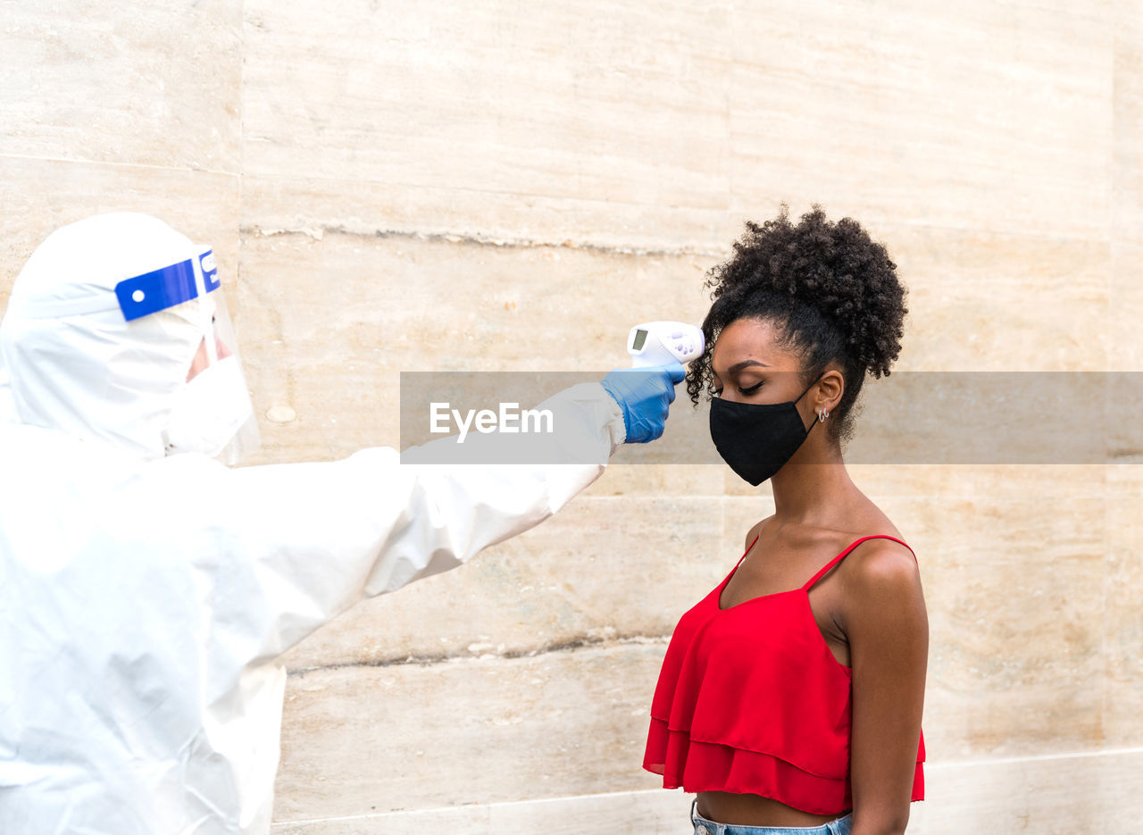 Woman checking temparature of girl standing against wall