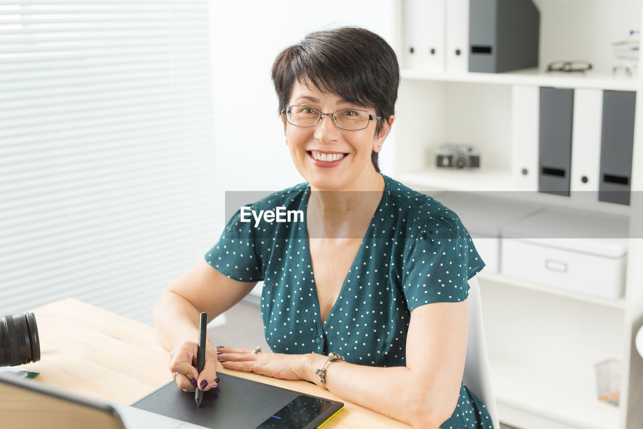 PORTRAIT OF A SMILING YOUNG WOMAN STANDING AGAINST TABLE