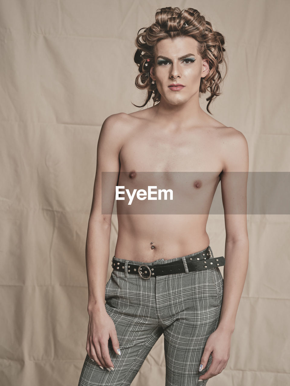 Shirtless slim androgynous male model with elegant curly hairdo and makeup looking at camera against beige fabric background