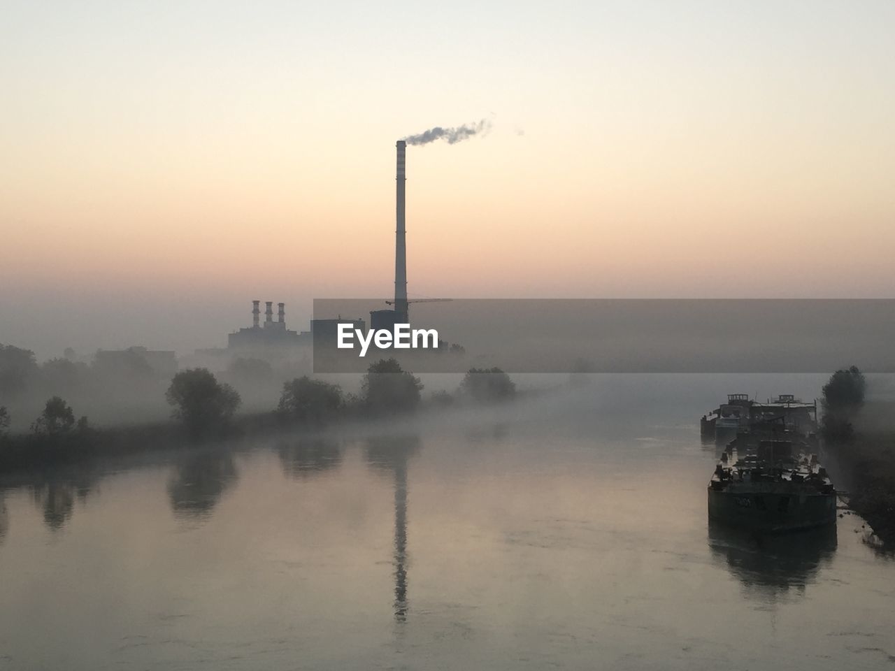 Smoke emitting from factory by river during foggy weather