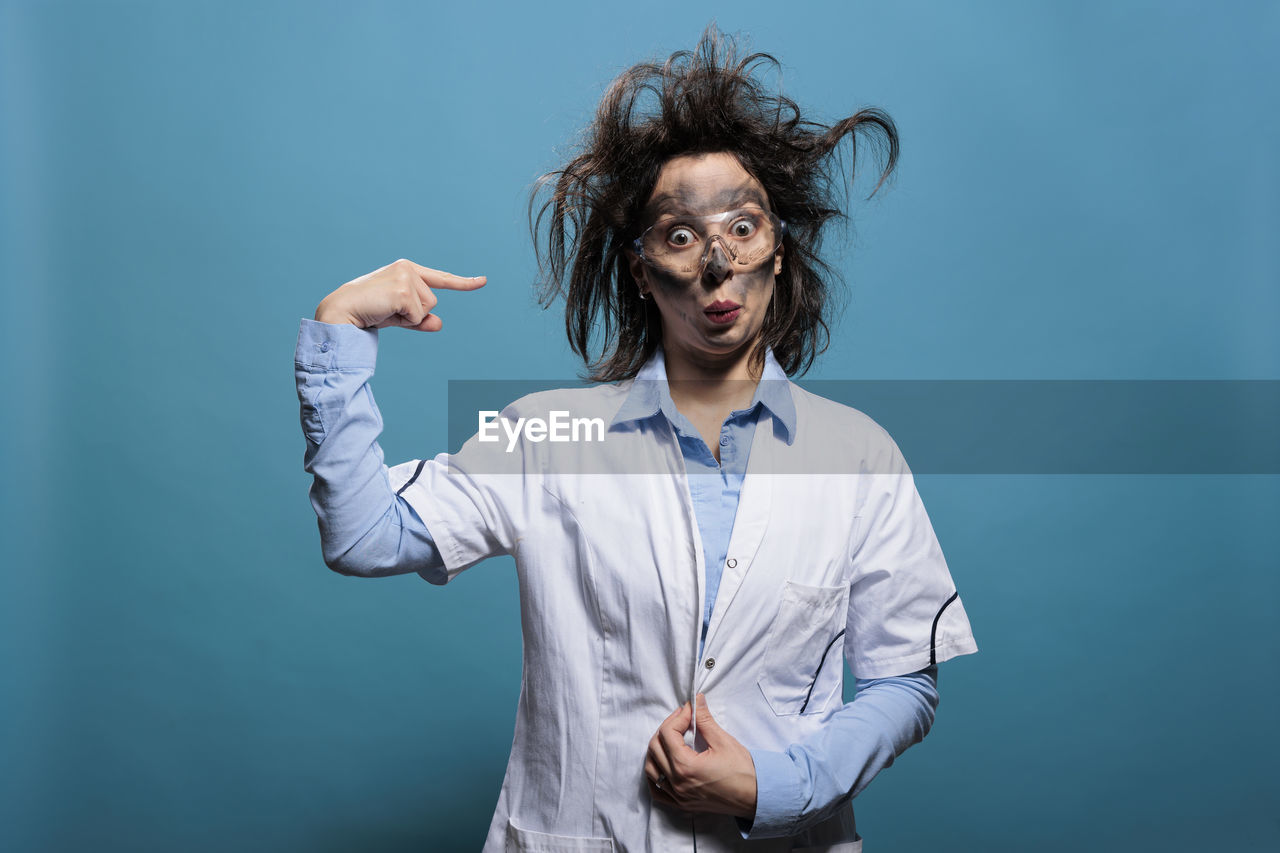 one person, studio shot, adult, blue, colored background, blue background, indoors, emotion, portrait, waist up, young adult, person, clothing, men, human face, photo shoot, standing, hairstyle, facial expression, front view, button down shirt, fun, casual clothing, negative emotion, looking at camera, gesturing, brown hair, business