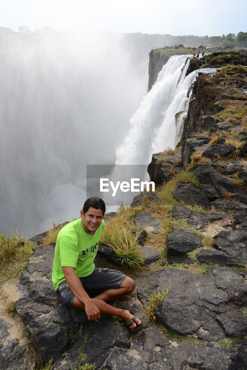 Man sitting on cliff against waterfall