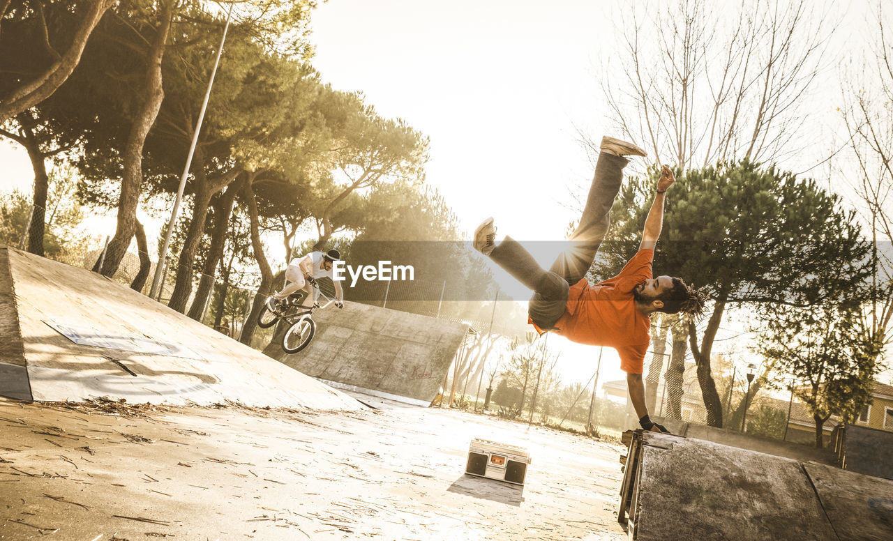 Man dancing with male friend riding bicycle at skateboard park during sunset