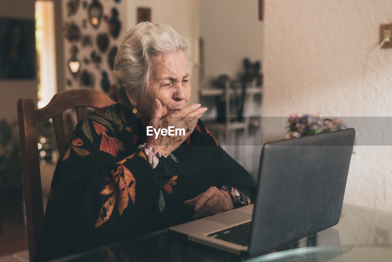 Positive elderly female wearing warm clothes sitting at table and sending air kiss while talking by video call using laptop