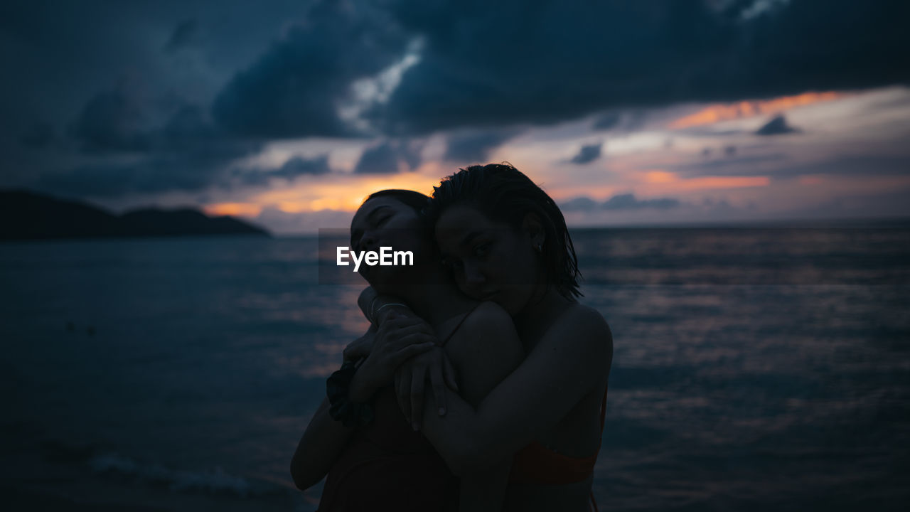 REAR VIEW OF COUPLE STANDING AT SEA DURING SUNSET