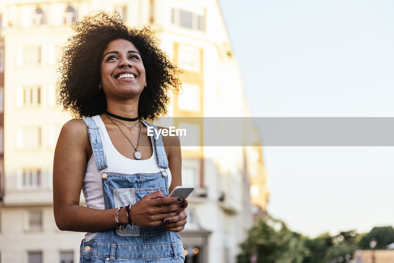 Smiling woman with phone standing against building