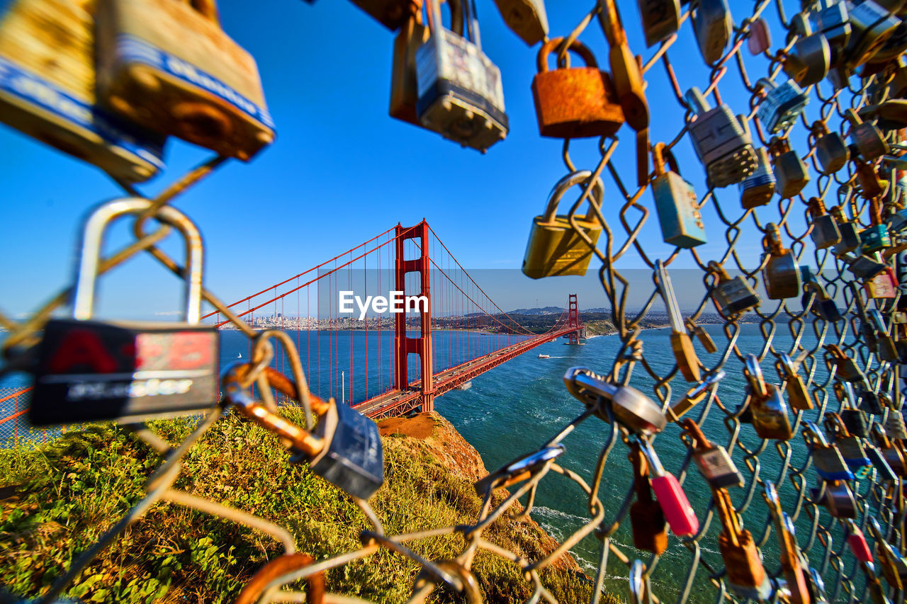 bridge, lock, padlock, sky, water, architecture, nature, built structure, metal, travel destinations, security, hanging, protection, transportation, no people, love lock, travel, fence, day, tourism, outdoors, suspension bridge, river, city, railing, positive emotion, amusement park, emotion, clear sky, blue, love, sunlight, chainlink fence