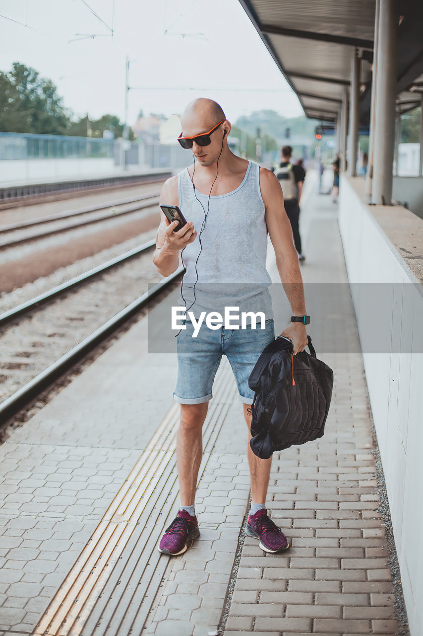 Young man in a t-shirt on the platform waiting for a train using mobile phone. man by train station 